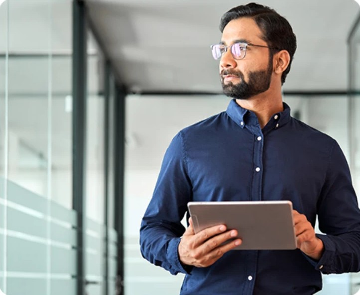 A man in glasses holding a tablet in a glass-walled office